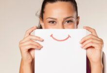 A woman holds a piece of paper over her face that has a smile printed on it. She stands in front of a white background.