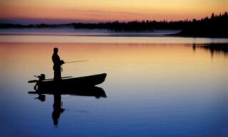 A silhouette of a man fishing at sunrise in a small boat on a lake. The sky and water are blue, orange, and purple.