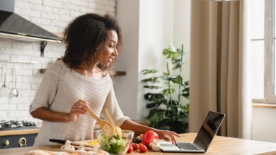 A woman standing in a kitchen mixing salad ingredients and chopping vegetables while looking at her laptop.