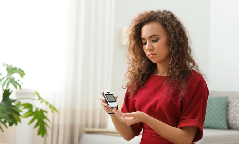 A brown-haired woman in a red shirt using a digital black and white glucometer to prick her finger at home.