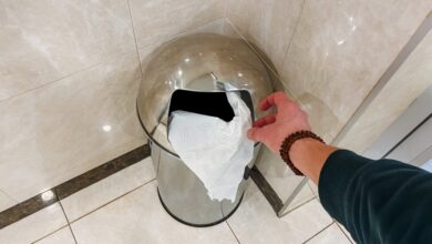 A person's hand tosses a piece of paper into a stainless steel trash can that sits in the corner of a tiled bathroom.