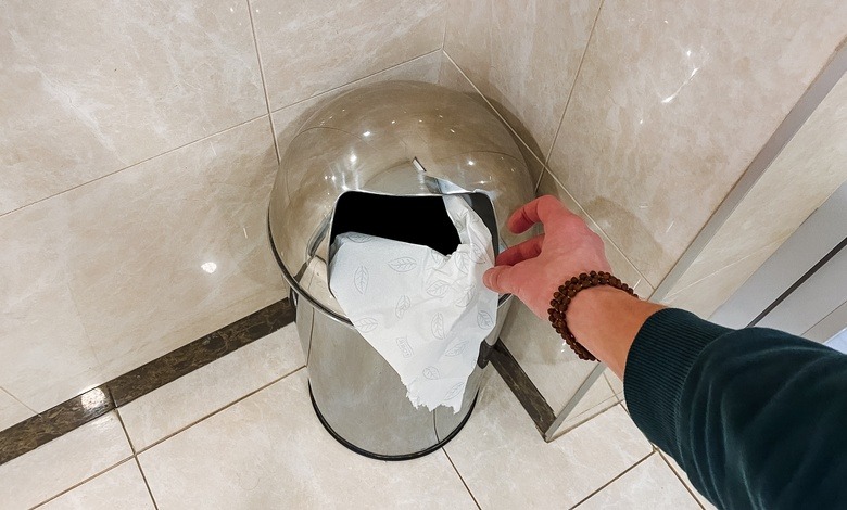 A person's hand tosses a piece of paper into a stainless steel trash can that sits in the corner of a tiled bathroom.