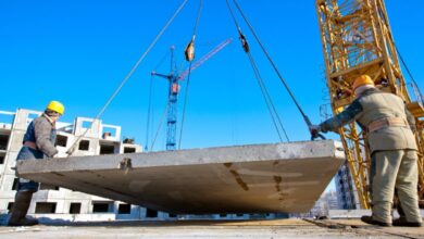 Two construction workers in hard hats positioning a concrete slab attached to wires on a building site.