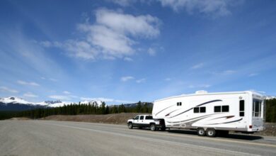 A white truck hauling a large white RV on an open road with trees and snowcapped mountains in the distance.