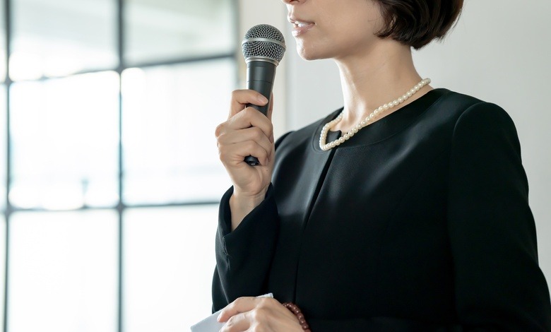 A woman in a black funeral dress and white pearls speaks into a microphone while holding a handkerchief in her hand.