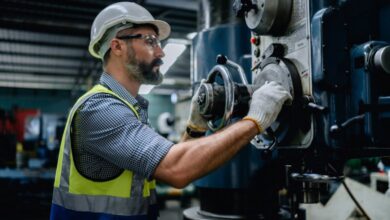 A worker wearing a safety vest, helmet, protective glasses, and gloves checks a handle on an industrial machine.