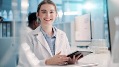 A young smiling woman in a white lab coat sits at a desk in a medical laboratory and holds a tablet.