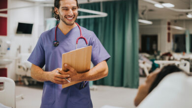 A smiling nurse standing beside a patient in a hospital bed. The nurse is holding a clipboard.