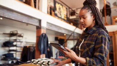 A long-haired young woman in a plaid shirt examining a silver tablet inside a two-story clothing store.