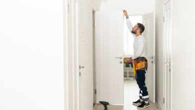 A handyman wearing a white shirt and a toolbelt installs a white wooden door in a bright, naturally lit hallway.