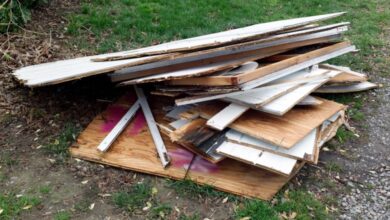 A large pile of scrap wood lies on patchy grass. Some of the wood is brown while other pieces are white.