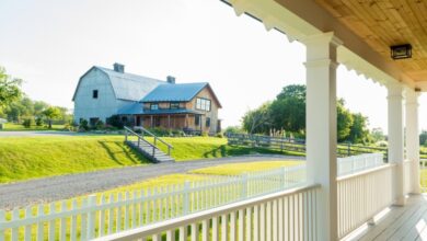 The view from a porch with white railings. A barn-style house with a gravel driveway sits in the distance.