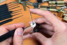 Close-up of a pair of hands trying to pick a cylinder lock with a lockpick and a tension wrench. Other tools lie nearby.