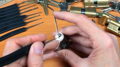 Close-up of a pair of hands trying to pick a cylinder lock with a lockpick and a tension wrench. Other tools lie nearby.
