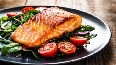 A large piece of salmon with mixed vegetables, including spinach and tomatoes, on a round black plate.