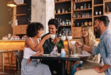 Two men and two women sit at a square table in a coffee shop, smiling and laughing while holding white mugs.