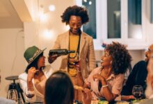 A group of seven people at a nighttime get-together in a warmly lit home. The host pours wine for a seated guest at a table.