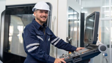 An engineer wearing a white hard hat and protective uniform smiles and uses a CNC machine control panel.