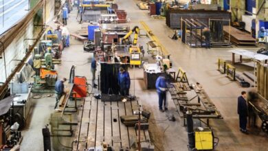 A large manufacturing shop floor with several metalworking machines, workbenches, and workers welding and assembling components.