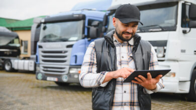 A man wearing a plaid shirt, black vest, and cap holding a tablet while standing in front of semi-trucks in a lot.